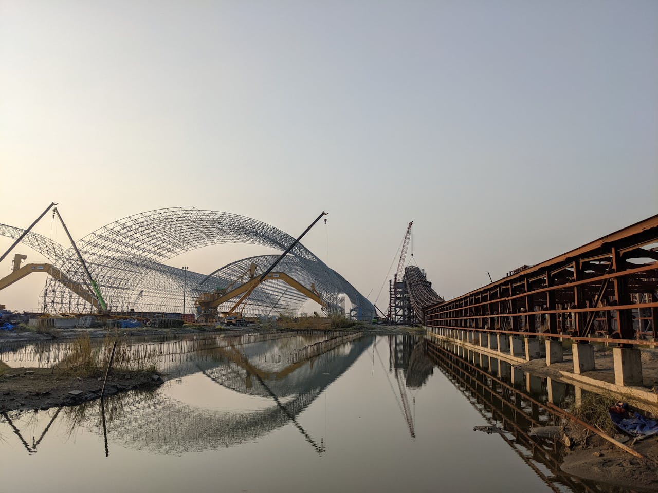 Home A picturesque industrial construction site with cranes at sunset, reflecting in calm water.